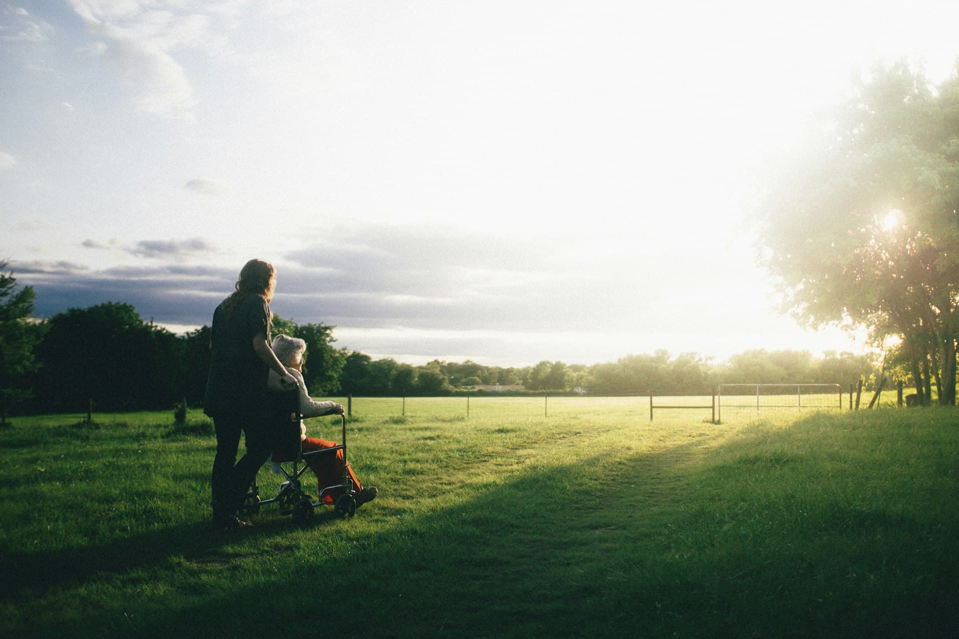 Caregiver walking with elderly woman in wheelchair outdoors at sunset